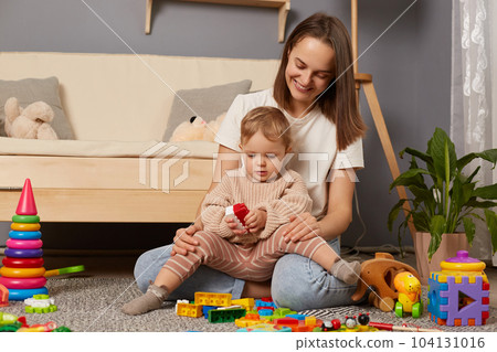 Image of smiling mother and child playing on the floor in the nursery with building a tower of colored blocks. Educational activities of children with parents at home. Image of smiling mother and child playing on the floor in the nursery with building a tower of colored blocks. Educational activities of children with parents at home. 104131016