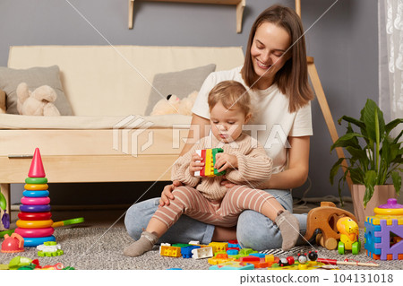 Horizontal shot of smiling dark haired woman in white t shirt sitting on floor with her baby daughter and playing with colorful blocks, spending free time for early development lesson. Horizontal shot of smiling dark haired woman in white t shirt sitting on floor with her baby daughter and playing with colorful blocks, spending free time for early development lesson. 104131018