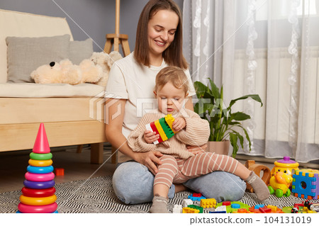 Happy positive young mother and cute infant daughter playing with colorful blocks, sitting on carpet at home, playing early developing games, expressing happiness. Happy positive young mother and cute infant daughter playing with colorful blocks, sitting on carpet at home, playing early developing games, expressing happiness. 104131019