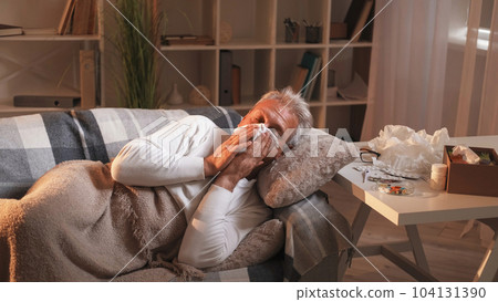 Runny nose. Home flu. Suffering man sneezing hard flu symptoms laying on sofa under plaid in light shadow room interior. 104131390
