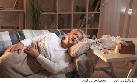 Fever symptoms. Home disease. Virus infection. Suffering man catching cold laying on sofa under plaid in light shadow room interior. 104131391