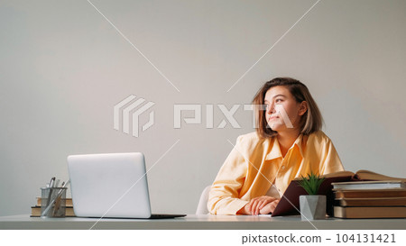 Laptop studying. Pensive freelancer. Remote workplace. Thoughtful girl working online distance job sitting at table against gray wall with empty space. 104131421