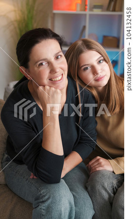 Family portrait. Happy relationship. Smiling mother teenage daughter enjoying spending time together sitting couch in light home interior. 104132458