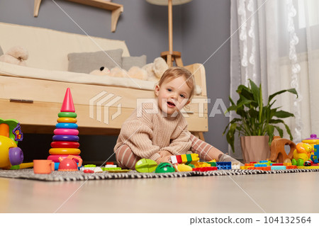 Portrait of cute smiling female kid wearing beige jumper sitting on floor surrounded with colorful toys, looking at camera, playing at home, smiling happily. Portrait of cute smiling female kid wearing beige jumper sitting on floor surrounded with colorful toys, looking at camera, playing at home, smiling happily. 104132564