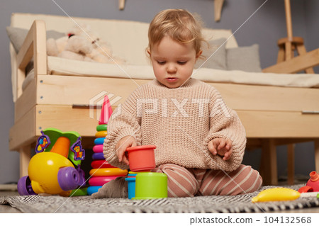 Portrait of little charming baby girl wearing beige sweater playing with colorful plastic toys, sitting on the floor, having fun and. Early learning. Development toys. Portrait of little charming baby girl wearing beige sweater playing with colorful plastic toys, sitting on the floor, having fun and. Early learning. Development toys. 104132568
