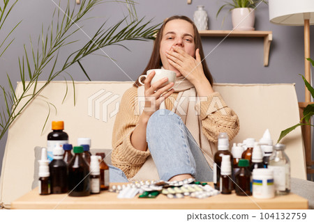 Portrait of sick sleepy woman wearing sweater and wrapped in scarf sitting on sofa at home, drinking hot tea from white cup, yawning, covering mouth with palm. Portrait of sick sleepy woman wearing sweater and wrapped in scarf sitting on sofa at home, drinking hot tea from white cup, yawning, covering mouth with palm. 104132759