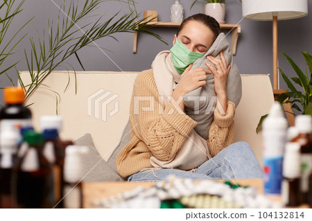 Indoor shot of sleepy tired Caucasian young adult woman wearing warm sweater, scarf and protective mask sitting on sofa in home interior among pills, holding pillow in hands and having nap. 104132814
