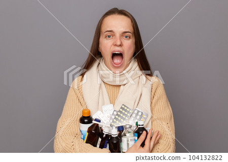 Portrait of panicked shocked caucasian woman wearing jumper and scarf holding pills, standing isolated over grey background, being seriously ill, screaming loud with despair. Portrait of panicked shocked caucasian woman wearing jumper and scarf holding pills, standing isolated over grey background, being seriously ill, screaming loud with despair. 104132822
