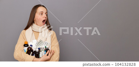 Portrait of surprised shocked caucasian woman wearing jumper and scarf holding pills, looking away at advertisement area, screaming from astonishment, standing isolated over grey background. 104132828