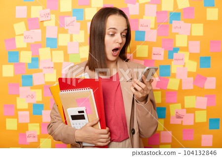 Shocked Caucasian attractive woman with brown hair wearing beige jacket holding paper folder and mobile phone, looking at display with open mouth, posing isolated over yellow background. Shocked Caucasian attractive woman with brown hair wearing beige jacket holding paper folder and mobile phone, looking at display with open mouth, posing isolated over yellow background. 104132846