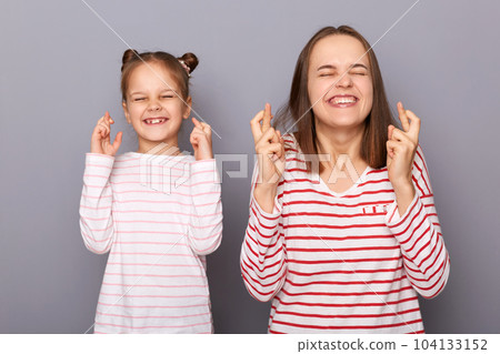 Indoor shot of smiling satisfied mother with daughter wearing casual clothing isolated over gray background, keeping finger closed, making wish, hopes for good luck. 104133152
