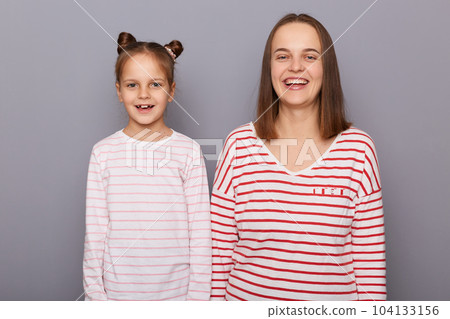 Horizontal shot of adorable beautiful woman and little girl with hair buns wearing casual clothes standing isolated over gray background, hearing funny joke, laughing. 104133156