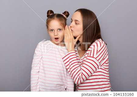 Indoor shot of mother whispering secret to her amazed with daughter who stands with open mouth, family wearing casual clothing isolated over gray background. Indoor shot of mother whispering secret to her amazed with daughter who stands with open mouth, family wearing casual clothing isolated over gray background. 104133160