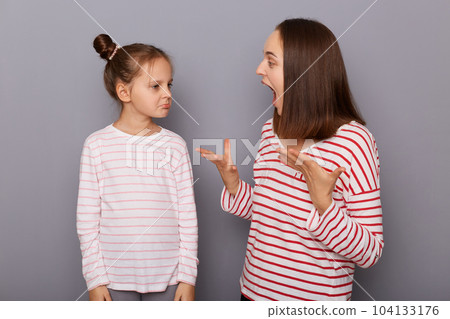 Portrait of irritated angry mother and sad daughter wearing casual clothes standing isolated over gray background, woman arguing with her kid, screaming with raised arms. 104133176