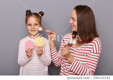 Portrait of cute positive woman and little girl with hair buns wearing casual clothes standing isolated over gray background, kid holding sponges, mommy applying powder with brush. 104133181