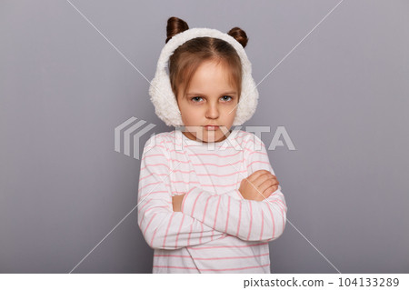 Image of little girl wearing fur earmuffs and striped shirt standing isolated over gray background, being offended, keeps arms folded, looking at cameras with pout lips. 104133289