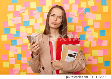 Portrait of sad upset disappointed woman wearing beige jacket standing against yellow wall with colorful memo cards, holding paper folders ans calculator, using cell phone, reading bad news. 104133369