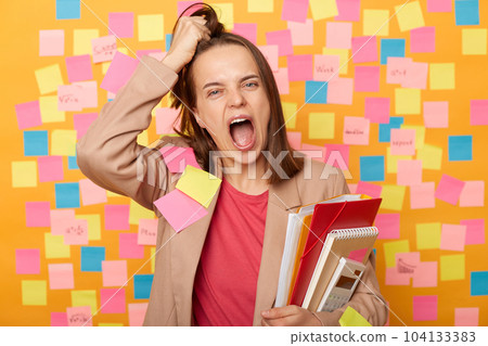 Photo of crazy tired young adult woman holding papers, posing against yellow wall with colorful stickers, shouting very loud, being exhausted, feels fatigue from work or studying, rips hair on head. 104133383
