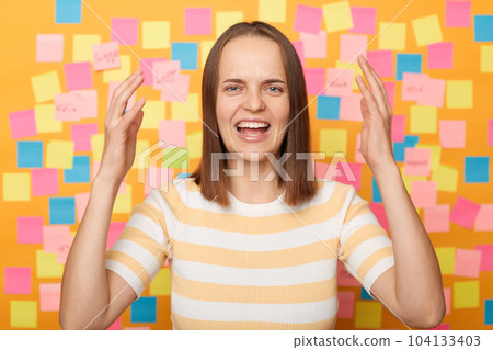 Photo of shocked sad despair brown haired woman wearing T-shirt, having serious problems, raised arms and screaming, posing against colorful sticky notes on yellow wall. Photo of shocked sad despair brown haired woman wearing T-shirt, having serious problems, raised arms and screaming, posing against colorful sticky notes on yellow wall. 104133403