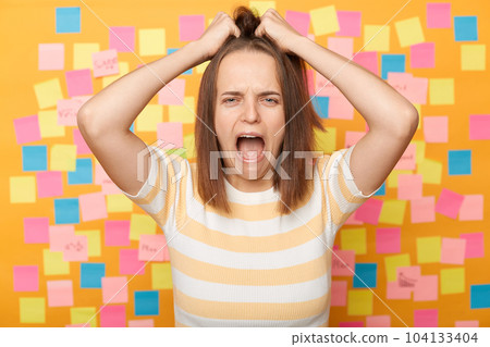 Indoor shot of irritated depressed woman with brown hair standing isolated over yellow background with stickers, screaming loud, pulling her hair, keeps mouth open. 104133404