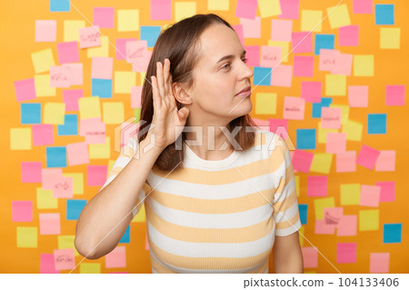 Portrait of curious woman wearing striped T-shirt posing over sticky notes to write reminder on yellow wall, standing with hand near ear, listening whispering. 104133406