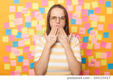 Indoor shot of shocked surprised beautiful Caucasian woman wearing striped T-shirt and glasses posing against yellow wall with colorful memo cards, sees something astonished, covered mouth with hands. Indoor shot of shocked surprised beautiful Caucasian woman wearing striped T-shirt and glasses posing against yellow wall with colorful memo cards, sees something astonished, covered mouth with hands. 104133417