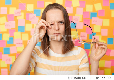 Stressed tired woman wearing casual t shirt standing isolated over yellow background with memo cards, rubbing eyes, putting off glasses, being tired work long hours. 104133422