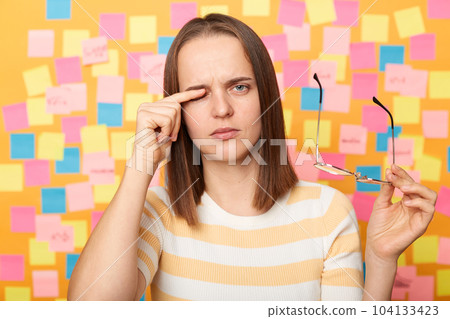 Horizontal shot of depressed sad tired exhausted woman wearing striped T-shirt, taking off glasses, rubbing eyes, feels pain, standing against yellow wall covered with stickers. 104133423