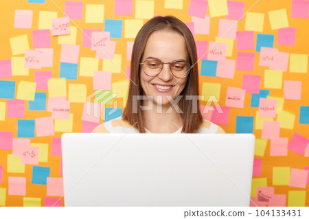 Horizontal shot of satisfied beautiful young adult woman wearing T-shirt posing against yellow paper wall with adhesive notes, sitting in front of white laptop, working online. Horizontal shot of satisfied beautiful young adult woman wearing T-shirt posing against yellow paper wall with adhesive notes, sitting in front of white laptop, working online. 104133431