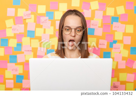 Indoor shot of shocked astonished woman in glasses with brown hair working on laptop against yellow wall with memo cards, looking monitor with open mouth, works online. Indoor shot of shocked astonished woman in glasses with brown hair working on laptop against yellow wall with memo cards, looking monitor with open mouth, works online. 104133432