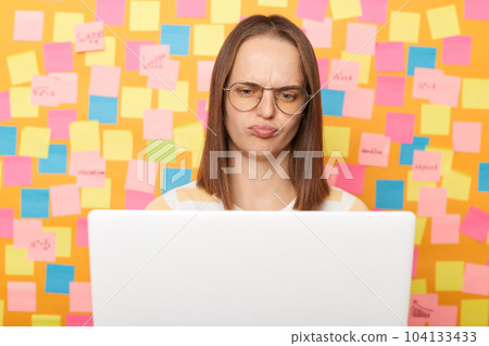 Photo of sad stresses tired brown haired woman wearing T-shirt posing against colorful sticky notes on yellow wall, working on laptop, looking at screen with sad expression. Photo of sad stresses tired brown haired woman wearing T-shirt posing against colorful sticky notes on yellow wall, working on laptop, looking at screen with sad expression. 104133433