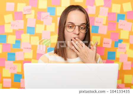 Portrait of amazed shocked surprised woman freelancer with dark hair wearing striped T-shirt, posing isolated over yellow background, working on laptop, covering mouth with palm. 104133435