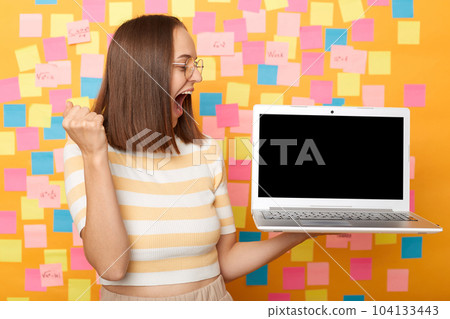 Photo of overjoyed happy joyful brown haired woman wearing T-shirt, looking at empty display for advertisement, clenched fist, celebrating, posing against colorful sticky notes on yellow wall. 104133443
