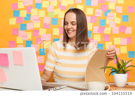 Image of adorable beautiful dark haired woman wearing striped t-shirt sitting at table, posing against memo cards on yellow wall, looking at notebook display and checking documents. 104133519