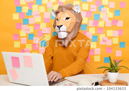 Indoor shot of anonymous man in lion mask wearing orange sweater sitting at workplace with laptop and typing on keyboard, working online, posing against yellow wall with stickers. Indoor shot of anonymous man in lion mask wearing orange sweater sitting at workplace with laptop and typing on keyboard, working online, posing against yellow wall with stickers. 104133633