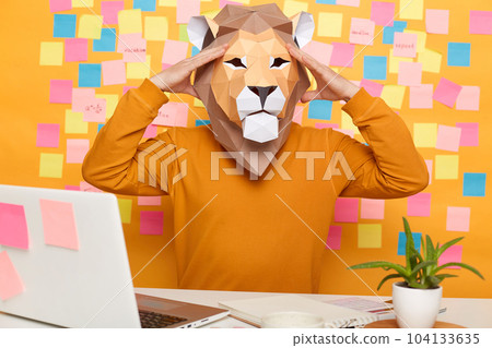 Image of tired sick unknown man in lion mask wearing orange jumper sitting at workplace with laptop against yellow wall with memo cards, having headache, works long hours. Image of tired sick unknown man in lion mask wearing orange jumper sitting at workplace with laptop against yellow wall with memo cards, having headache, works long hours. 104133635