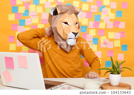 Indoor shot of sick tired unknown man in lion mask wearing orange jumper sitting at workplace with laptop against yellow wall with memo cards, feels pain in his back, massaging painful neck. Indoor shot of sick tired unknown man in lion mask wearing orange jumper sitting at workplace with laptop against yellow wall with memo cards, feels pain in his back, massaging painful neck. 104133643