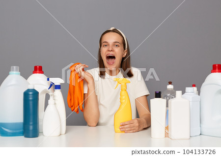 Image of amazed happy woman wears headband and casual t shirt, sits at workplace with different detergents, holding rubber gloves in hands, isolated over gray background. Image of amazed happy woman wears headband and casual t shirt, sits at workplace with different detergents, holding rubber gloves in hands, isolated over gray background. 104133726