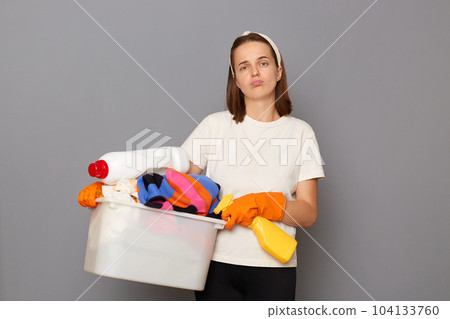 Portrait of sad tired woman wearing t shirt and hair band holding basin with clothing isolated over gray background, feels fatigue, being exhausted, doing lots work about the house, doing laundry. 104133760