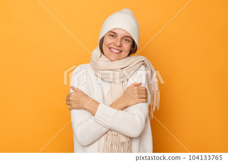 Portrait of self-satisfied egoistic cheerful woman wearing white sweater, hat and scarf posing isolated over yellow background, embracing herself and smiling with pleasure, feeling self-pride. Portrait of self-satisfied egoistic cheerful woman wearing white sweater, hat and scarf posing isolated over yellow background, embracing herself and smiling with pleasure, feeling self-pride. 104133765