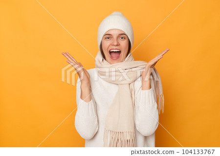 Photo of excited amazed satisfied woman wearing white sweater, scarf and cap, raised her arms, being happy to win, celebrating her success, standing isolated over yellow background. Photo of excited amazed satisfied woman wearing white sweater, scarf and cap, raised her arms, being happy to win, celebrating her success, standing isolated over yellow background. 104133767