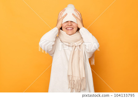 Indoor shot of smiling satisfied woman wearing sweater, scarf and hat, hiding her eyes with cap, smiling happily, having fun, feels shy, standing isolated over yellow background. 104133795