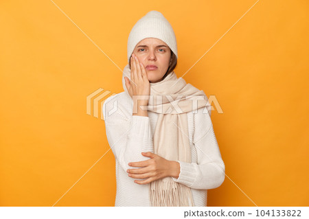 Indoor shot of sick ill unhealthy Caucasian woman wearing warm jumper, cap and scarf posing isolated over yellow background, feels terrible pain in her teeth. 104133822
