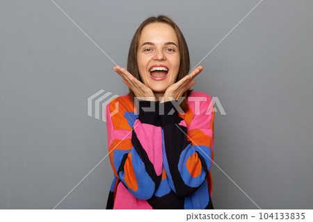 Portrait of extremely happy young adult woman wearing colorful sweater posing isolated over gray background, keeps palms under chin, screaming, celebrating her success. Portrait of extremely happy young adult woman wearing colorful sweater posing isolated over gray background, keeps palms under chin, screaming, celebrating her success. 104133835