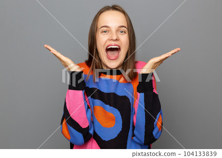 Indoor shot of carefree optimistic woman with brown hair wearing sweater standing isolated over gray background, raised her arms, yelling with overjoyed expression, hearing very good news, rejoice. 104133839
