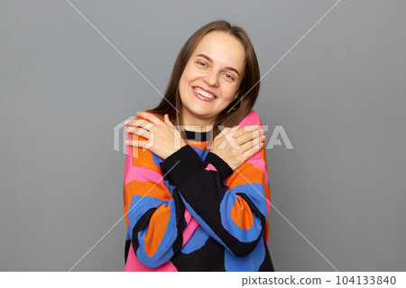 Portrait of pretty positive lovely woman with brown hair wearing colorful jumper, standing looking at camera with satisfied expression, hugging herself, posing isolated over gray background. 104133840