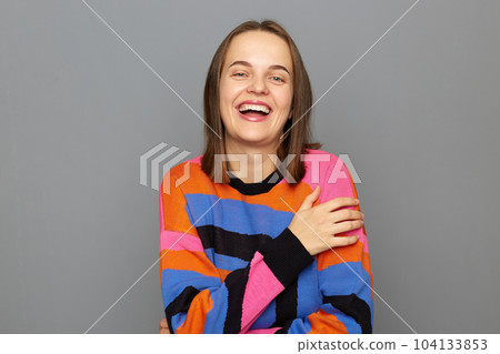 Indoor shot of laughing Caucasian woman wearing colorful sweater posing isolated over gray background, laughs out loud, looking at camera, hearing funny joke. Indoor shot of laughing Caucasian woman wearing colorful sweater posing isolated over gray background, laughs out loud, looking at camera, hearing funny joke. 104133853