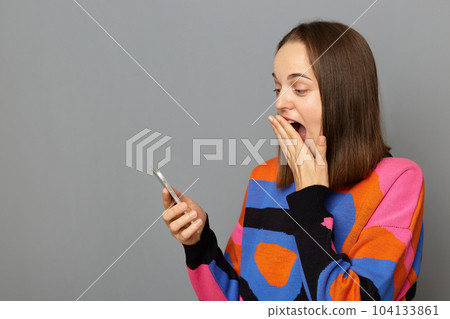 Image of excited woman with brown hair wearing jumper standing isolated over grey background, holding cell phone, reading surprised good news, copy space for advertisement. Image of excited woman with brown hair wearing jumper standing isolated over grey background, holding cell phone, reading surprised good news, copy space for advertisement. 104133861