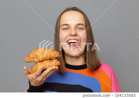 portrait of cheerful smiling Caucasian woman wearing sweater, holding in hands two croissants, looking at camera, sticking tongue out, posing isolated over gray background. portrait of cheerful smiling Caucasian woman wearing sweater, holding in hands two croissants, looking at camera, sticking tongue out, posing isolated over gray background. 104133883