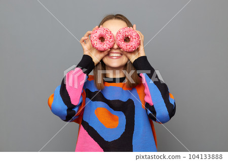 Smiling happy Caucasian woman wearing sweater posing isolated over gray background, keeps sweet donuts on eyes, having fun with tasty dessert. Dieting, junk food, weight loss. Smiling happy Caucasian woman wearing sweater posing isolated over gray background, keeps sweet donuts on eyes, having fun with tasty dessert. Dieting, junk food, weight loss. 104133888